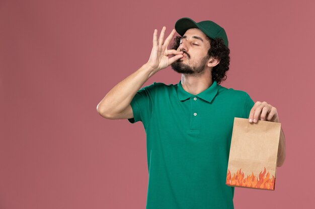 Mensajero masculino de vista frontal en uniforme verde y capa que sostiene el paquete de alimentos de papel sobre fondo rosa servicio de entrega uniforme