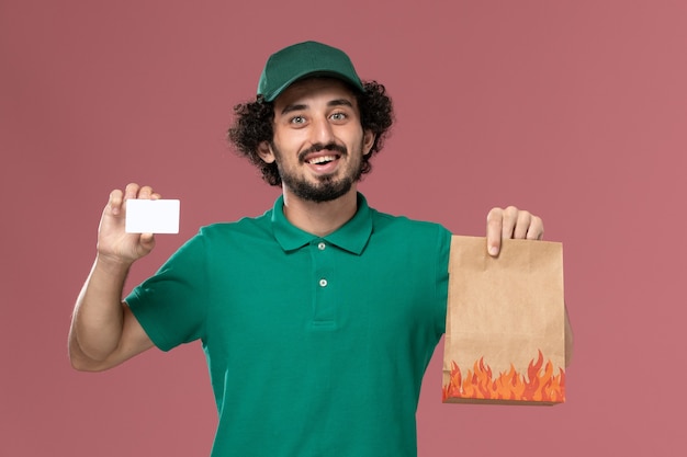 Mensajero masculino de vista frontal en uniforme verde y capa que sostiene la tarjeta y el paquete de alimentos en el trabajo de trabajo masculino de entrega uniforme de servicio de fondo rosa