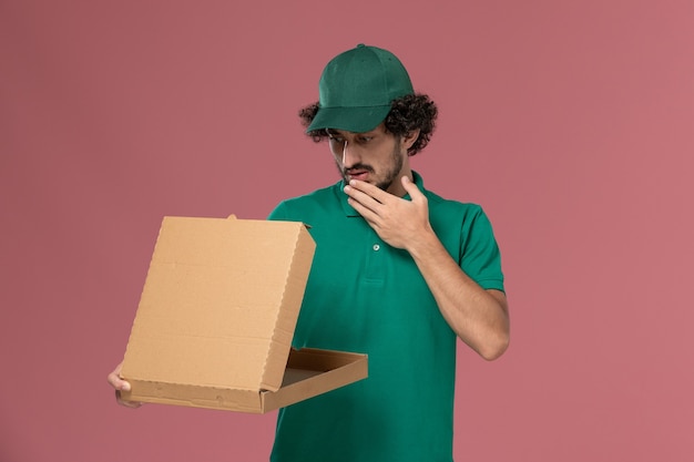 Mensajero masculino de vista frontal en uniforme verde y capa sosteniendo y abriendo la caja de comida de entrega en el fondo rosa Servicio de entrega uniforme