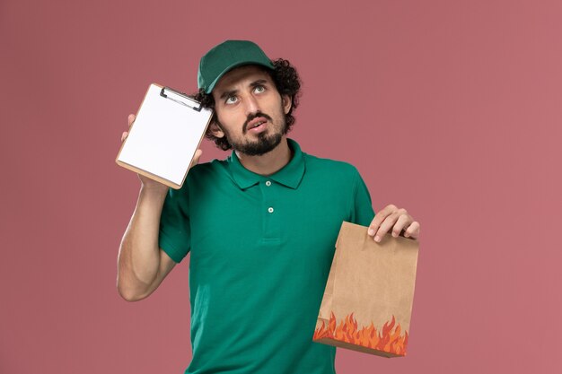 Mensajero masculino de vista frontal en uniforme verde y capa sosteniendo el bloc de notas y el paquete de alimentos pensando en fondo rosa servicio uniforme de entrega masculino