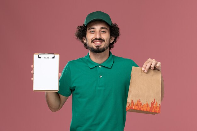 Mensajero masculino de vista frontal en uniforme verde y capa sosteniendo el bloc de notas y paquete de alimentos sobre fondo rosa servicio uniforme de entrega masculino