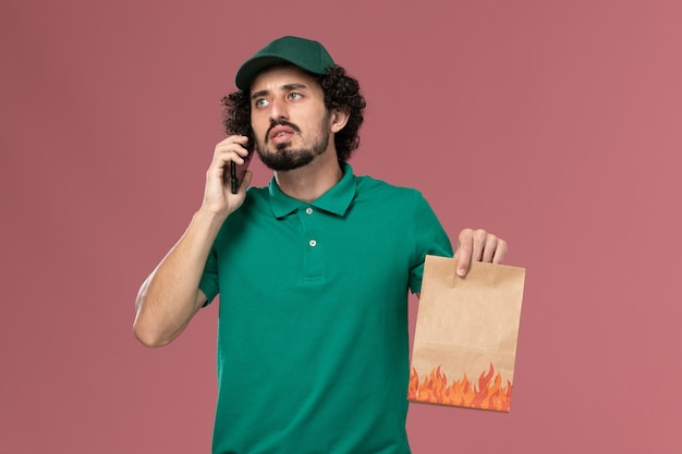 Mensajero masculino de vista frontal en uniforme verde y capa sosteniendo el paquete de alimentos y hablando por teléfono sobre fondo rosa servicio uniforme entrega trabajo masculino