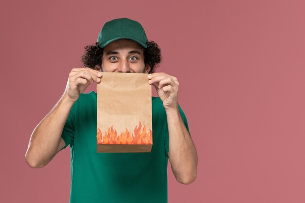 Mensajero masculino de vista frontal en uniforme verde y capa sosteniendo el paquete de alimentos de papel sobre fondo rosa servicio de entrega uniforme masculino