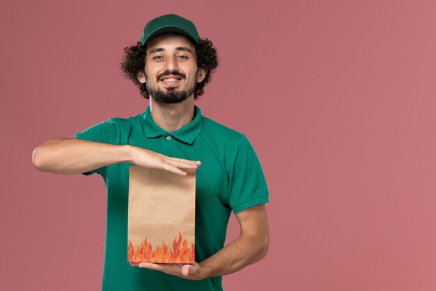 Mensajero masculino de vista frontal en uniforme verde y capa sosteniendo el paquete de alimentos de papel y sonriendo sobre fondo rosa trabajador de servicio entrega uniforme