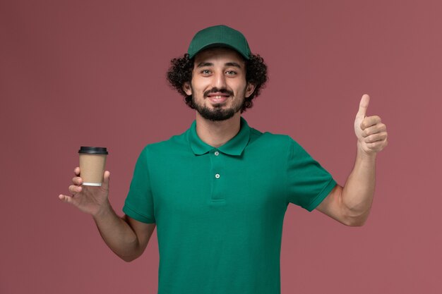 Mensajero masculino de vista frontal en uniforme verde y capa sosteniendo la taza de café de entrega en la empresa de trabajo de servicio de entrega uniforme de fondo rosa