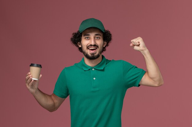 Mensajero masculino de vista frontal en uniforme verde y capa sosteniendo la taza de café de entrega en el fondo rosa servicio de entrega uniforme trabajo trabajador trabajo empresa