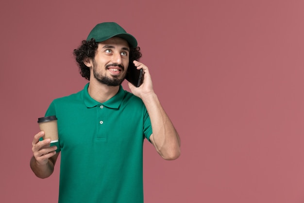 Mensajero masculino de vista frontal en uniforme verde y capa sosteniendo la taza de café de entrega y hablando por teléfono en el servicio de trabajo de entrega uniforme de escritorio rosa