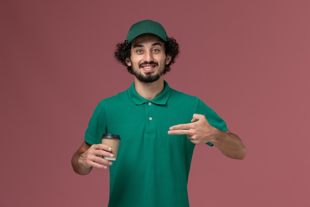 Mensajero masculino de vista frontal en uniforme verde y capa sosteniendo la taza de café de entrega posando en el trabajo de servicio de entrega uniforme de fondo rosa