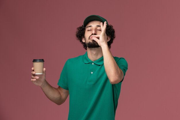 Mensajero masculino de vista frontal en uniforme verde y capa sosteniendo la taza de café de entrega sobre fondo rosa servicio de entrega uniforme trabajo empresa masculina