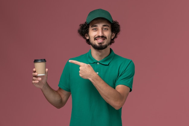 Mensajero masculino de vista frontal en uniforme verde y capa sosteniendo la taza de café de entrega con una sonrisa en el trabajo de servicio de entrega uniforme de fondo rosa