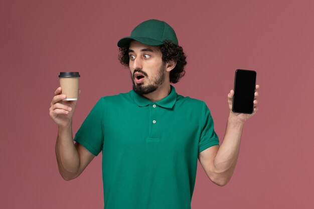 Mensajero masculino de vista frontal en uniforme verde y capa sosteniendo la taza de café de entrega con el teléfono en el servicio de trabajo de entrega uniforme de fondo rosa