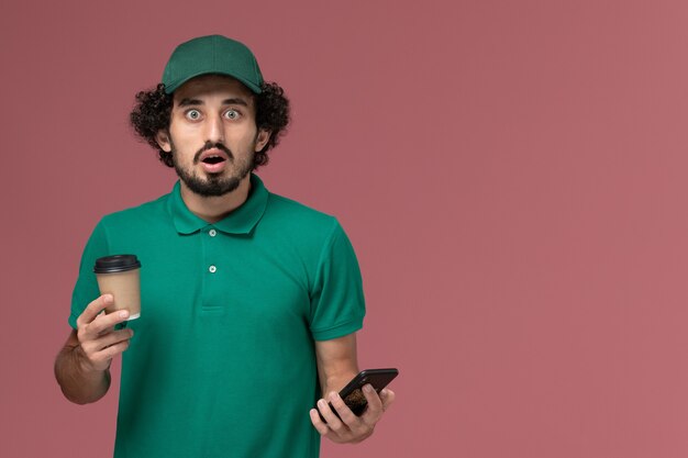 Mensajero masculino de vista frontal en uniforme verde y capa sosteniendo la taza de café de entrega usando su teléfono en el servicio de trabajo de entrega uniforme de fondo rosa