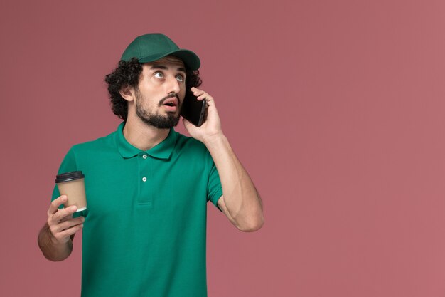 Mensajero masculino de vista frontal en uniforme verde y capa sosteniendo la taza de café y hablando por teléfono en el servicio de entrega uniforme de fondo rosa