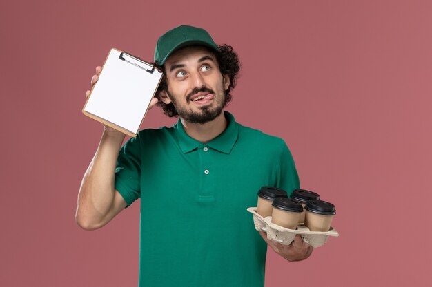 Mensajero masculino de vista frontal en uniforme verde y capa sosteniendo tazas de café con bloc de notas pensando en el trabajo de entrega uniforme de servicio de fondo rosa