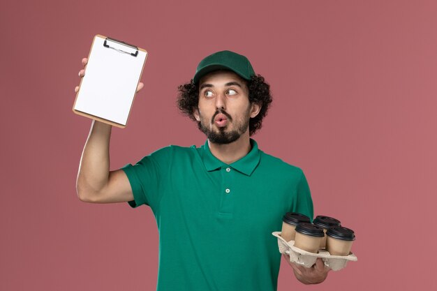 Mensajero masculino de vista frontal en uniforme verde y capa sosteniendo tazas de café con bloc de notas en el trabajo de entrega uniforme de servicio de fondo rosa masculino