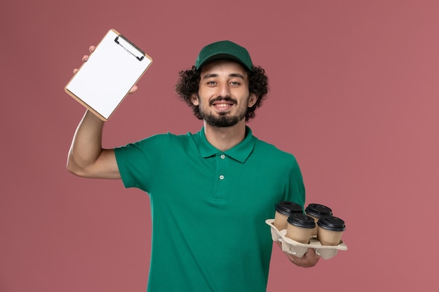 Mensajero masculino de vista frontal en uniforme verde y capa sosteniendo tazas de café con bloc de notas en el trabajo de trabajador de entrega uniforme de servicio de fondo rosa