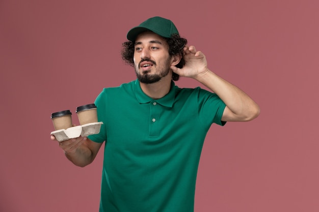 Mensajero masculino de vista frontal en uniforme verde y capa sosteniendo tazas de café de entrega en el fondo rosa servicio trabajo trabajador masculino entrega uniforme
