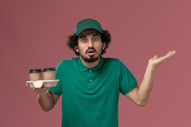 Mensajero masculino de vista frontal en uniforme verde y capa sosteniendo tazas de café de entrega sobre fondo rosa trabajo de entrega uniforme trabajo de trabajador masculino