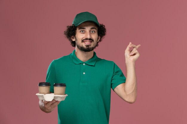 Mensajero masculino de vista frontal en uniforme verde y capa sosteniendo tazas de café de entrega con sonrisa en servicio de escritorio rosa trabajo trabajador masculino entrega uniforme