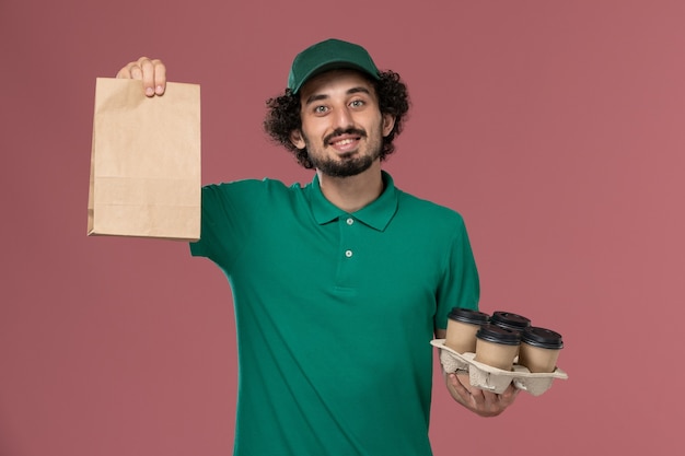 Mensajero masculino de vista frontal en uniforme verde y capa sosteniendo tazas de café con paquete de alimentos sobre fondo rosa servicio de entrega uniforme trabajo trabajador masculino