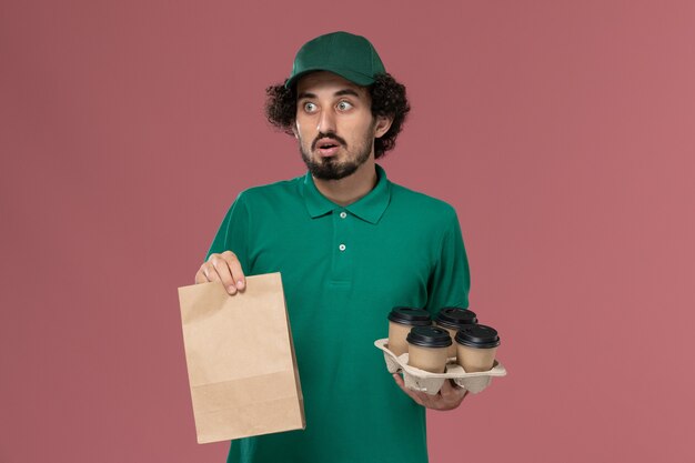 Mensajero masculino de vista frontal en uniforme verde y capa sosteniendo tazas de café con paquete de comida sobre fondo rosa servicio de entrega uniforme trabajador masculino