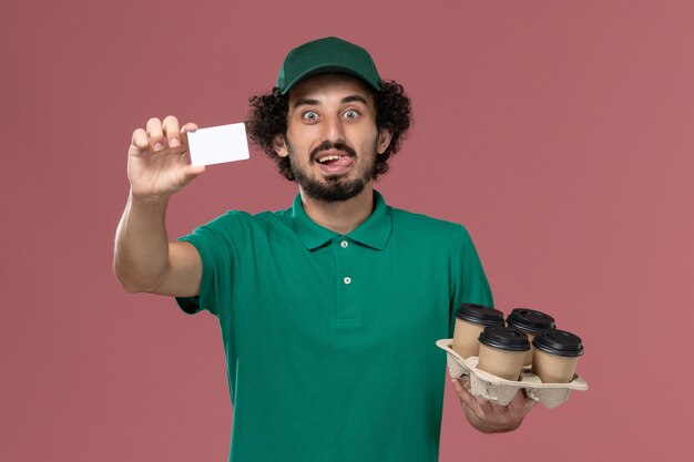 Mensajero masculino de vista frontal en uniforme verde y capa sosteniendo tazas de café con tarjeta sobre fondo rosa servicio de entrega uniforme masculino