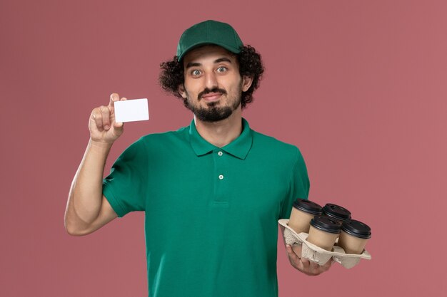 Mensajero masculino de vista frontal en uniforme verde y capa sosteniendo tazas de café con tarjeta sobre fondo rosa servicio de entrega uniforme masculino