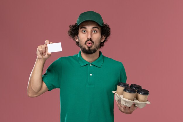 Mensajero masculino de vista frontal en uniforme verde y capa sosteniendo tazas de café con tarjeta en el trabajador de trabajo de entrega uniforme de servicio de fondo rosa