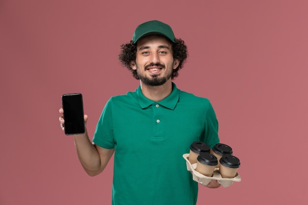 Foto gratuita mensajero masculino de vista frontal en uniforme verde y capa sosteniendo tazas de café con teléfono en el trabajo de entrega uniforme de servicio de fondo rosa claro
