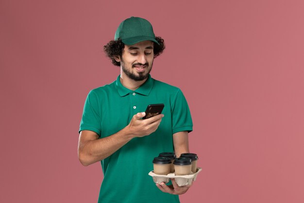 Mensajero masculino de vista frontal en uniforme verde y capa sosteniendo tazas de café tomando fotos de ellos sobre fondo rosa servicio uniforme entrega trabajo trabajador masculino