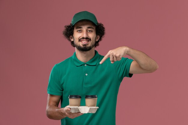 Mensajero masculino de vista frontal en uniforme verde y capa sosteniendo tazas de café en el trabajo de servicio de entrega uniforme de fondo rosa