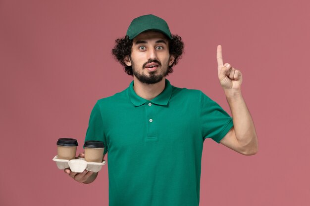 Mensajero masculino de vista frontal en uniforme verde y capa sosteniendo tazas de café en el trabajo de trabajador de servicio de entrega uniforme de fondo rosa