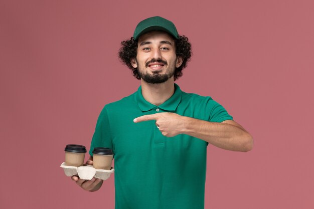 Mensajero masculino de vista frontal en uniforme verde y capa sosteniendo tazas de café en el trabajo de trabajador de servicio de entrega uniforme de fondo rosa