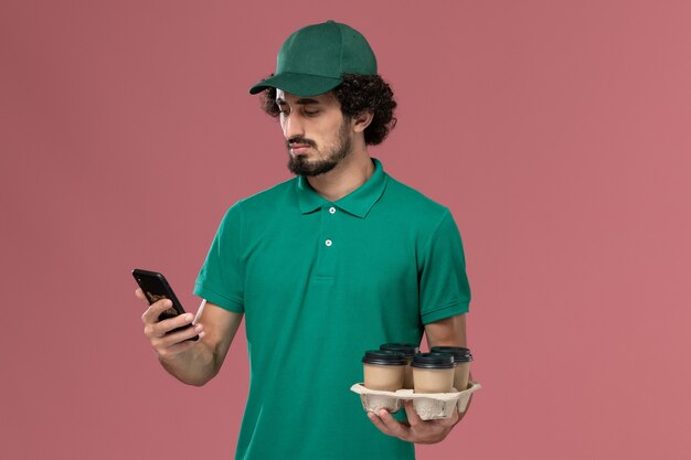 Mensajero masculino de vista frontal en uniforme verde y capa sosteniendo tazas de café usando su teléfono en el trabajo masculino de entrega uniforme de servicio de fondo rosa