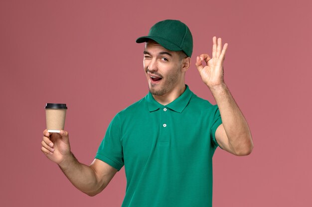 Mensajero masculino de vista frontal en uniforme verde guiñando un ojo y sosteniendo la taza de café sobre el fondo rosa