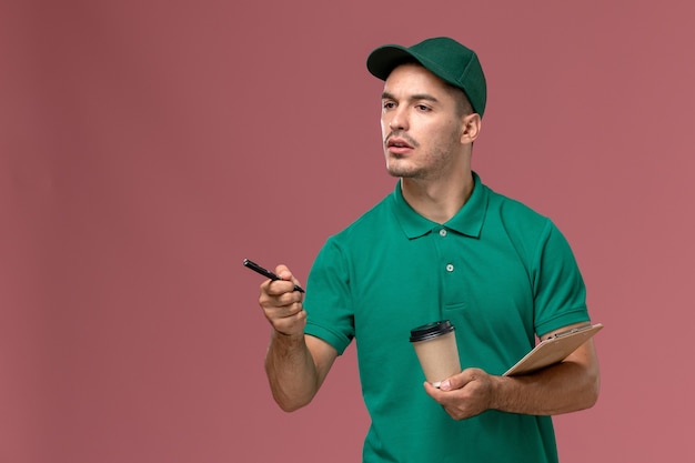 Mensajero masculino de la vista frontal en uniforme verde que sostiene el bloc de notas y el bolígrafo de la taza de café de entrega sobre fondo rosa claro