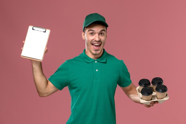 Mensajero masculino de la vista frontal en uniforme verde que sostiene el bloc de notas de tazas de café marrón sobre el fondo rosa