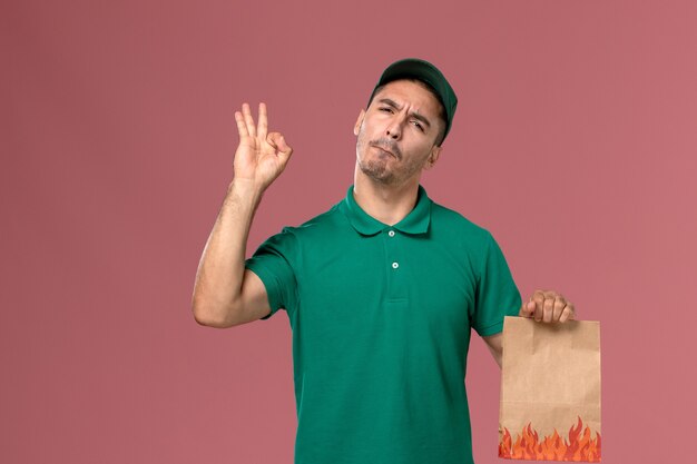 Mensajero masculino de la vista frontal en uniforme verde que sostiene el paquete de comida de papel en el fondo rosa claro