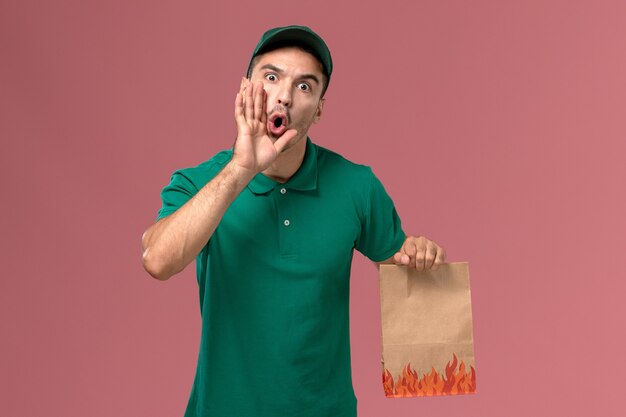 Mensajero masculino de la vista frontal en uniforme verde que sostiene el paquete de comida de papel en el fondo rosa claro