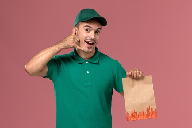 Mensajero masculino de la vista frontal en uniforme verde que sostiene el paquete de comida de papel en el fondo rosa claro