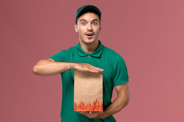 Mensajero masculino de la vista frontal en uniforme verde que sostiene el paquete de comida de papel en el fondo rosa