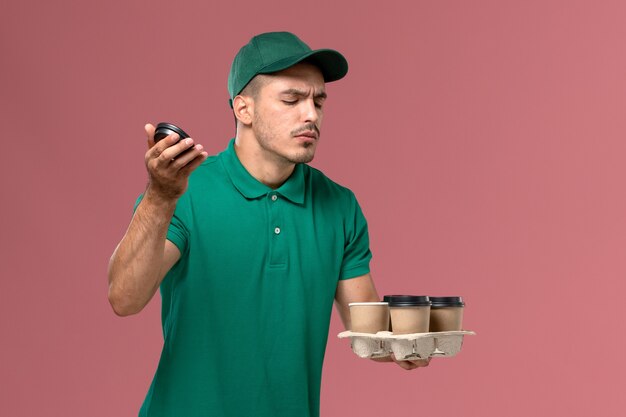 Mensajero masculino de la vista frontal en uniforme verde que sostiene las tazas de café de la entrega que huelen en el fondo rosado