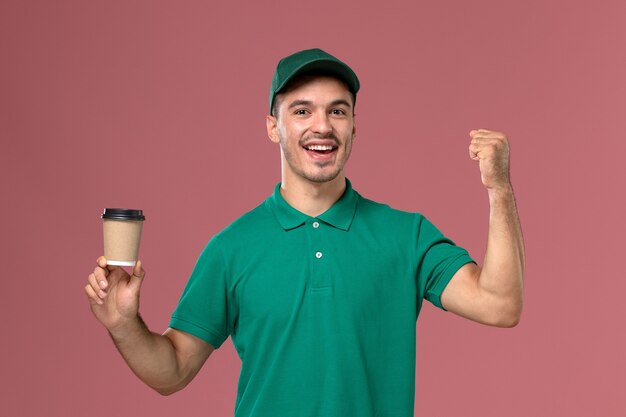 Mensajero masculino de vista frontal en uniforme verde regocijándose y sosteniendo la taza de café sobre el fondo rosa