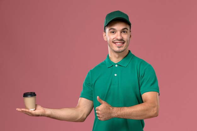 Mensajero masculino de vista frontal en uniforme verde sonriendo y sosteniendo la taza de café sobre fondo rosa claro