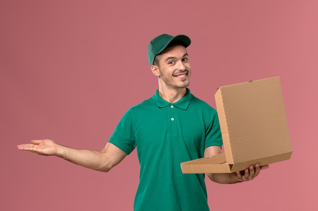 Mensajero masculino de vista frontal en uniforme verde sosteniendo y abriendo la caja de comida en el fondo rosa claro