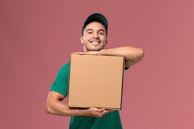 Mensajero masculino de vista frontal en uniforme verde sosteniendo la caja de comida y sonriendo en el escritorio rosa