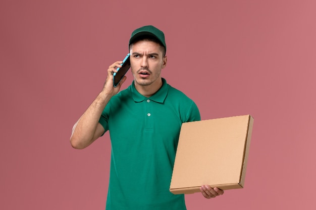 Mensajero masculino de vista frontal en uniforme verde sosteniendo la caja de comida y usando su teléfono en el escritorio rosa