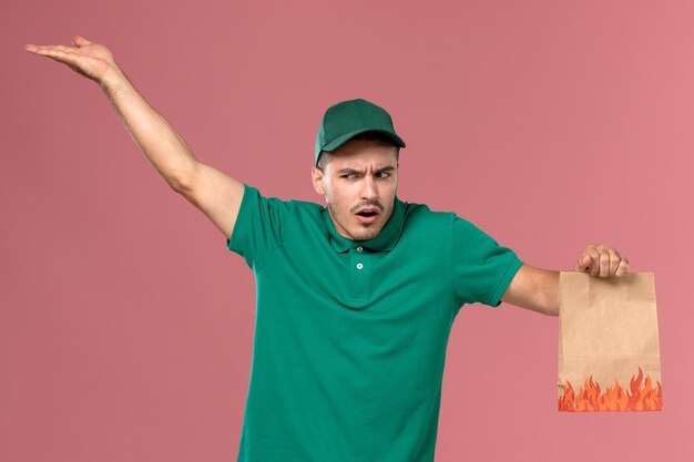 Mensajero masculino de vista frontal en uniforme verde sosteniendo el paquete de alimentos con expresión confusa en su rostro sobre fondo rosa claro