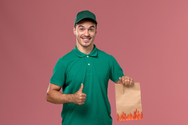 Mensajero masculino de vista frontal en uniforme verde sosteniendo el paquete de alimentos y sonriendo sobre el fondo rosa claro