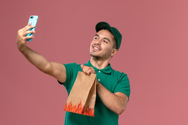 Mensajero masculino de vista frontal en uniforme verde sosteniendo el paquete de alimentos y tomando fotos con él en el fondo rosa